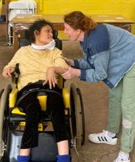 Two people are smiling at each other. One is in a wheelchair, the other is bending down to make eye contact. 